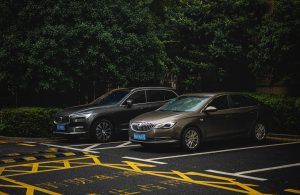 Two cars, an SUV and a sedan, parked side by side in a shady urban lot. Deux voitures, un SUV et une berline, garées côte à côte dans un parking urbain ombragé.