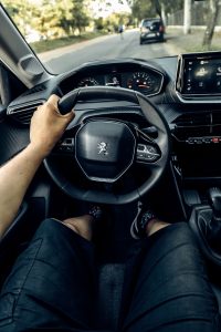 View of a person driving a Peugeot car, focused on the steering wheel and dashboard in a lush green setting. Vue d'une personne conduisant une voiture Peugeot, en mettant l'accent sur le volant et le tableau de bord dans un cadre verdoyant.